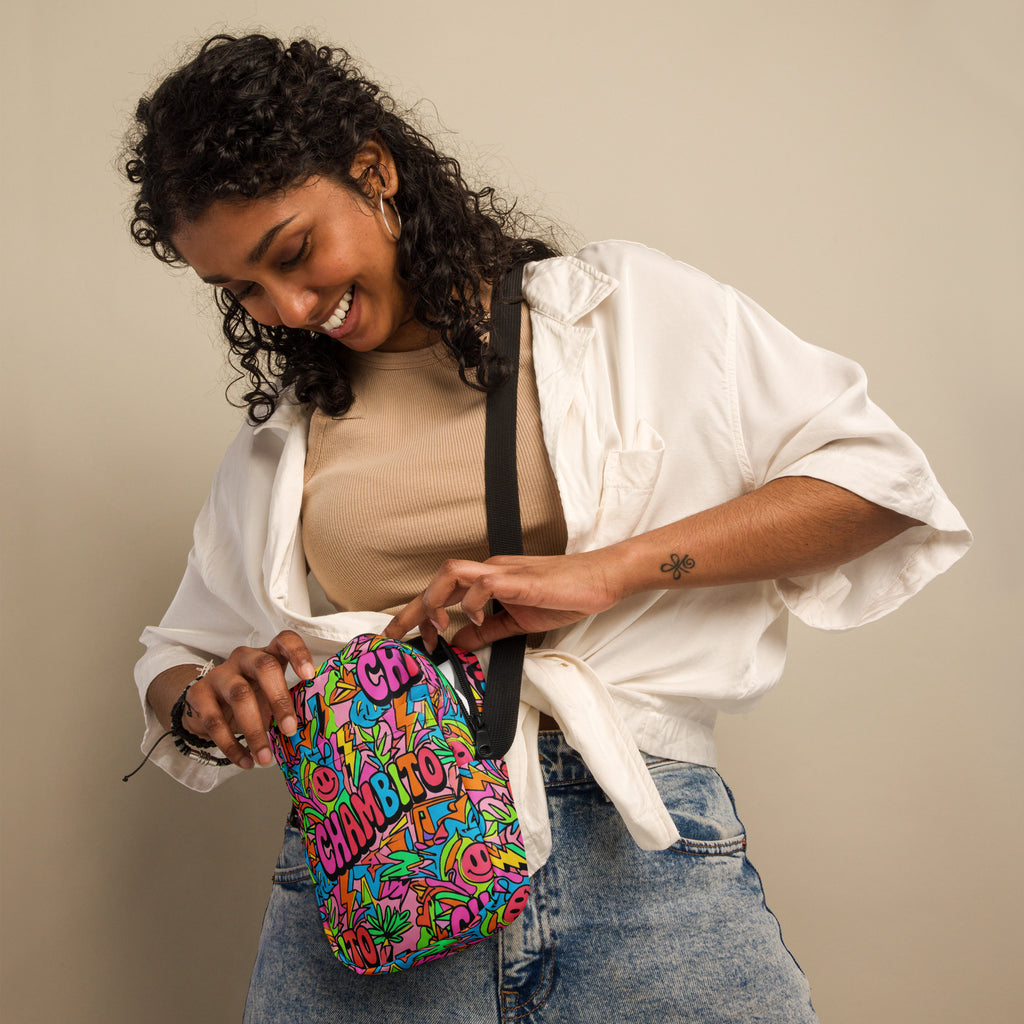 A woman wearing a white button-up shirt and blue jeans holds a colorful crossbody bag with a black strap. The bag features a vibrant, multicolored graffiti-style pattern with the words CHAMBITO